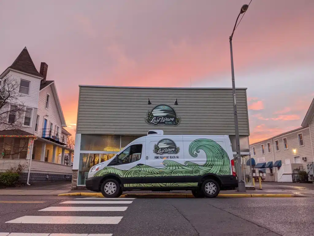 Van with ocean wave design parked outside Last Wave Brewery in Point Pleasant Beach, NJ, against a colorful sunset backdrop, highlighting dog-friendly outdoor seating area.