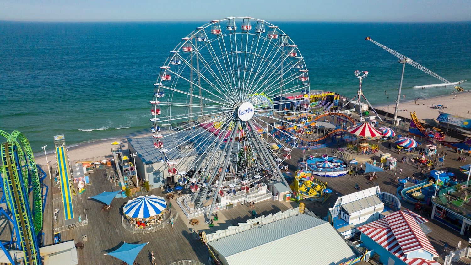 Ferris wheel at Casino Pier in Seaside Heights, with colorful amusement rides and beach in background, promoting the grand opening event on April 12th.