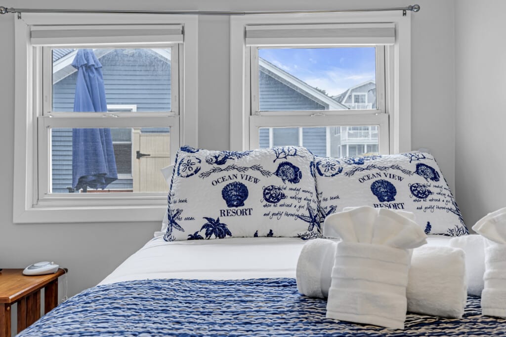 Cozy bedroom in a Jersey Shore vacation rental featuring "Ocean View Resort" decorative pillows, white towels, and blue patterned bedding, with windows showcasing a view of a blue umbrella and neighboring houses.