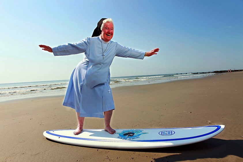 Nun in a surf outfit balancing on a surfboard at a sandy beach, showcasing a fun and unique surfing experience, reflecting the vibrant surf culture of Nun's Beach in Stone Harbor, New Jersey.