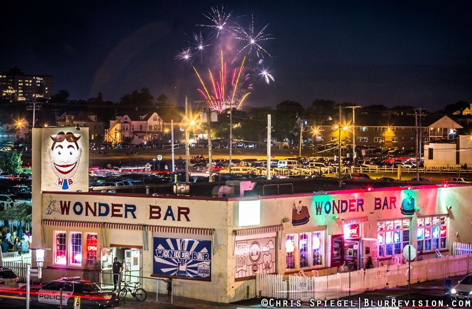 Fireworks illuminating the night sky above Wonder Bar, a dog-friendly venue in Asbury Park, featuring vibrant exterior lights and a lively atmosphere, surrounded by parked cars and buildings.