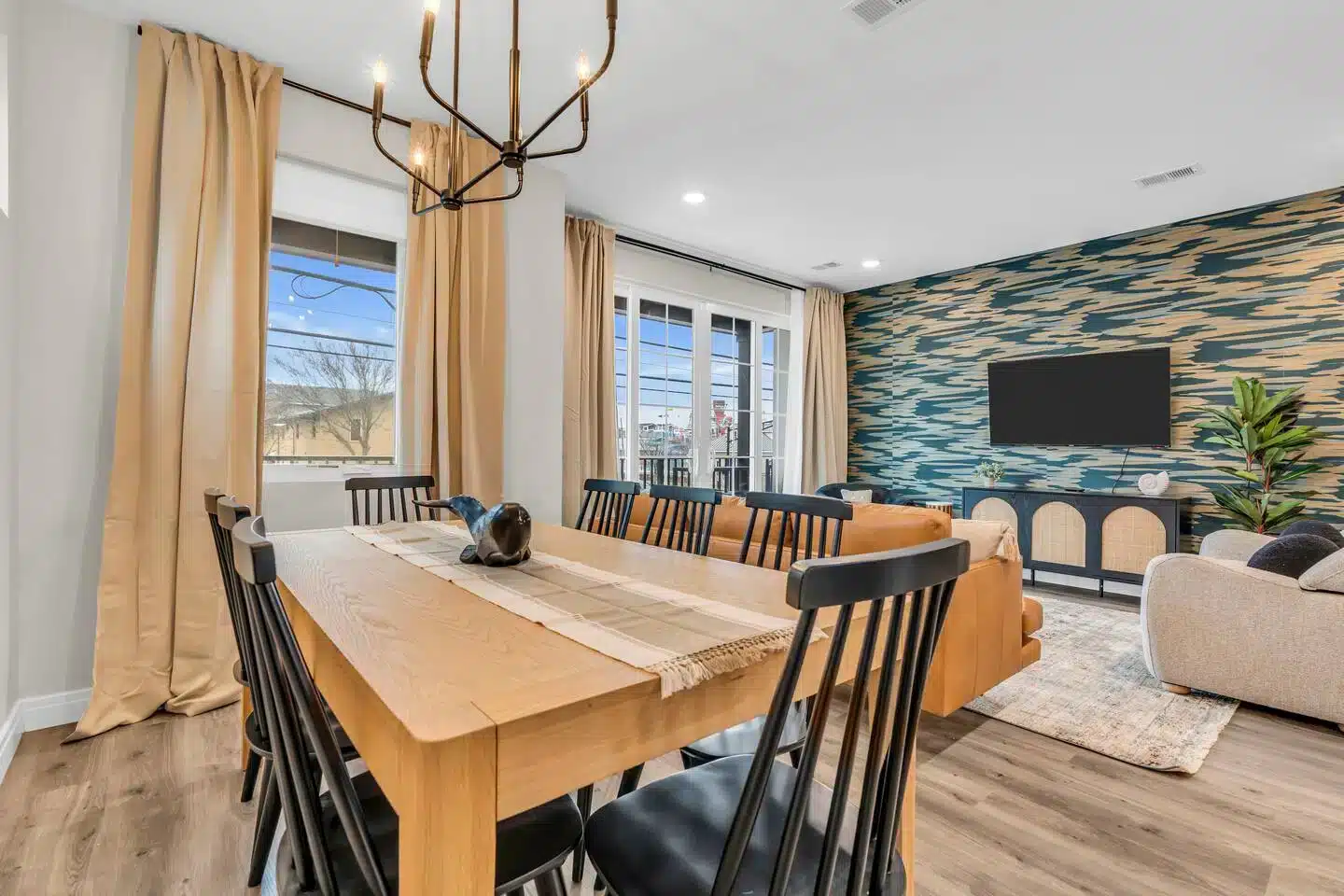 Modern dining area with wooden table and black chairs, featuring large windows with natural light, stylish decor, and a cozy living space visible in the background, emphasizing design for comfort and functionality in vacation rentals.