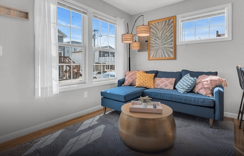 Cozy living room featuring a blue sectional sofa with colorful pillows, a gold accent table, and large windows allowing natural light, ideal for a family-friendly Jersey Shore vacation rental.