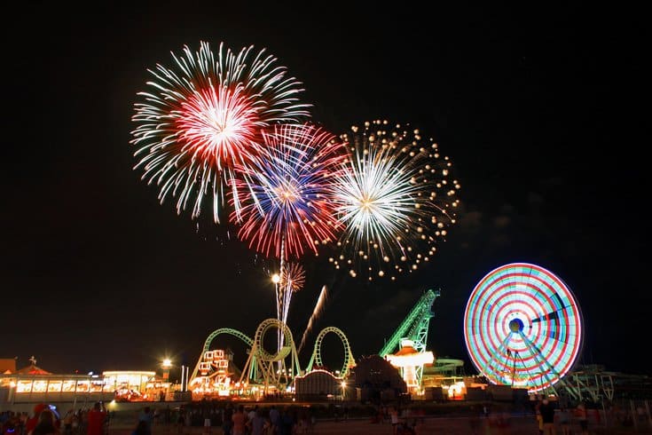 Fireworks display over amusement rides at night, colorful explosions illuminating the sky above a boardwalk with a Ferris wheel and roller coaster, capturing the festive atmosphere of summer nights at the Jersey Shore.