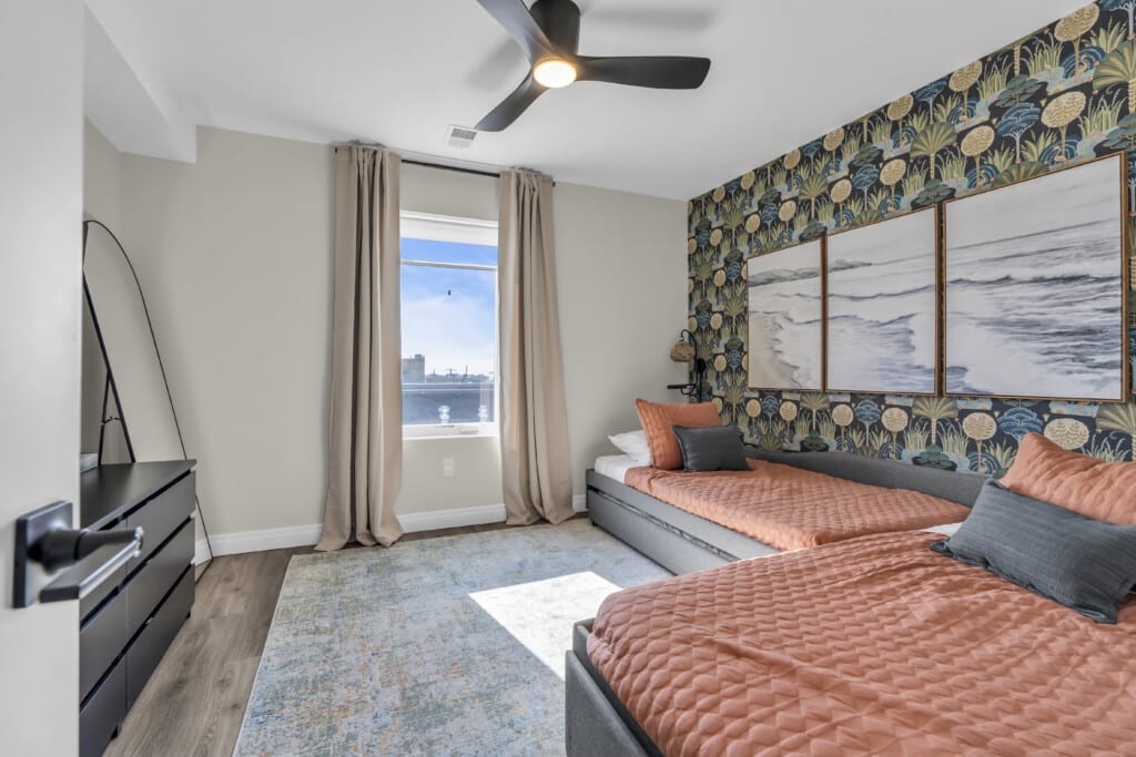 Cozy bedroom featuring two beds with coral bedding, a large window with ocean views, and beach-themed wall art, ideal for vacation rental appeal.