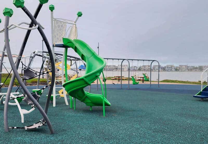 Modern green playground equipment with a slide and swings, overlooking a calm bay, ideal for family bonding and outdoor activities near vacation rentals.
