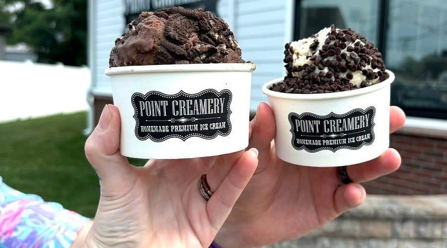 Two cups of homemade premium ice cream from Point Creamery, one with chocolate cookie dough and the other with vanilla topped with chocolate chips, held by a person against a blurred outdoor background.