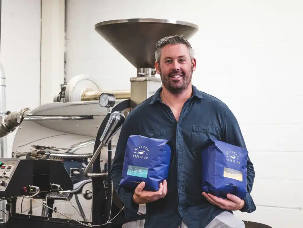 Man holding two bags of Offshore Coffee Co. coffee in a roasting facility, showcasing the brand's surfer-inspired aesthetic and commitment to quality brews, relevant to Jersey Shore coffee shop recommendations.