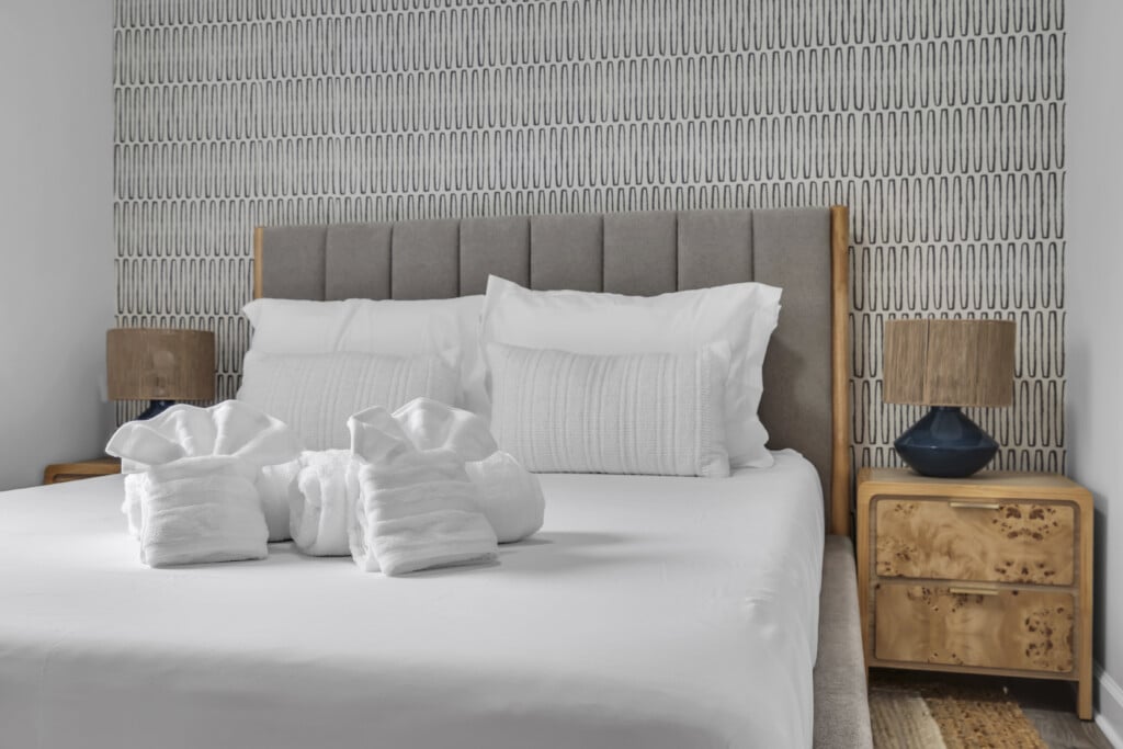 Modern bedroom with a neatly made bed featuring white linens and decorative towels, accented by a patterned wall and stylish bedside lamps, reflecting the inviting atmosphere of vacation rentals at the Jersey Shore.