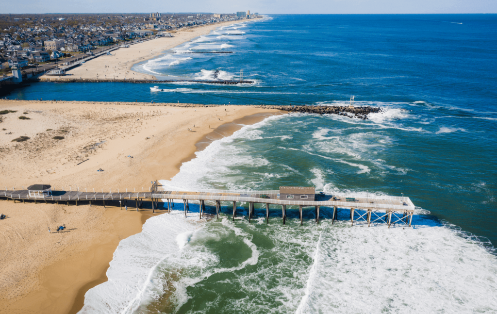 Aerial view of Belmar Beach, New Jersey, featuring a wooden pier extending into the ocean, sandy shoreline, and surfable waves, highlighting a popular surfing destination at the Jersey Shore.