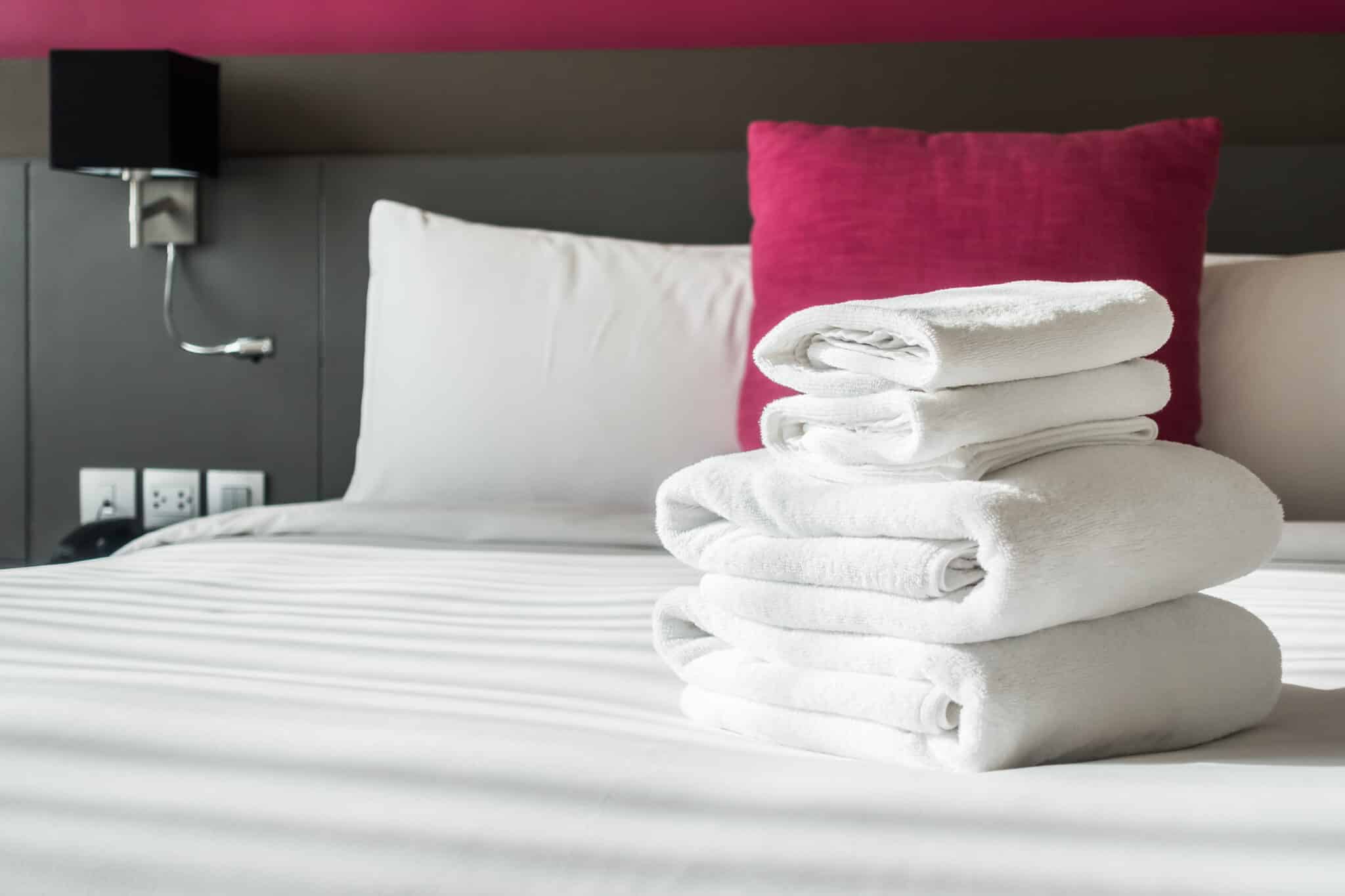 Folded white bath towels on a neatly made bed, with a pink accent pillow and bedside lamp, emphasizing essential packing items for a beach vacation rental at the Jersey Shore.