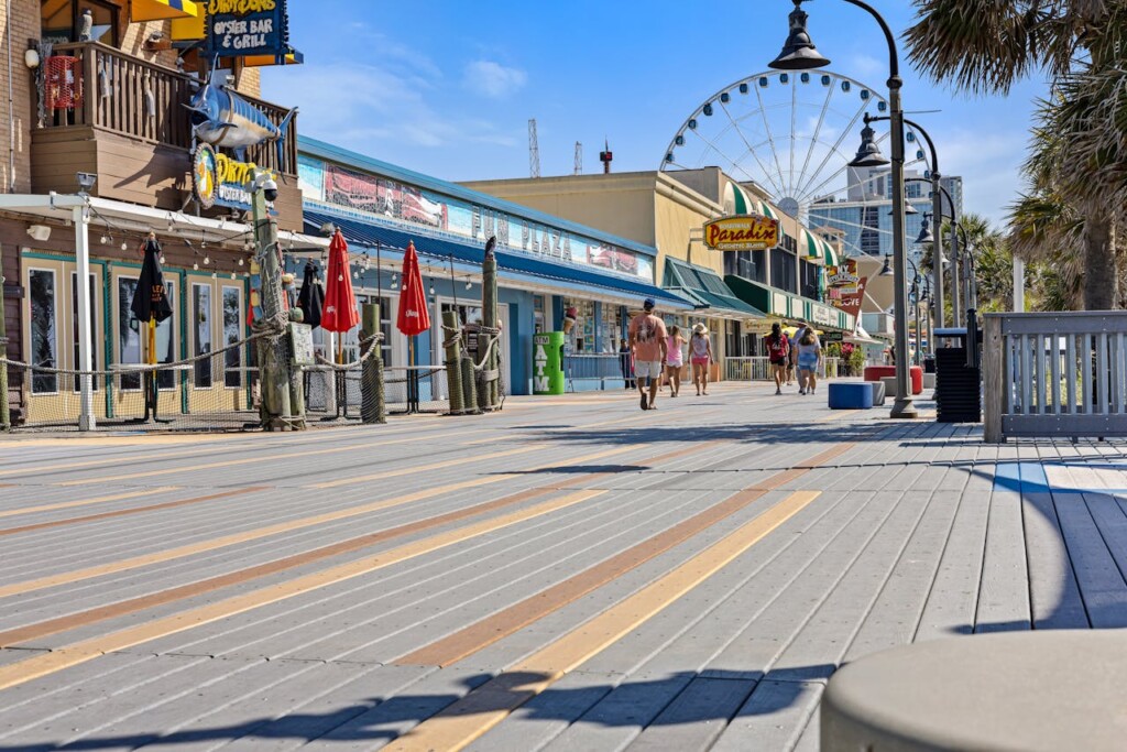 Boardwalk scene at the Jersey Shore featuring shops, diners, and a Ferris wheel, with visitors strolling along the wooden walkway under a clear blue sky.