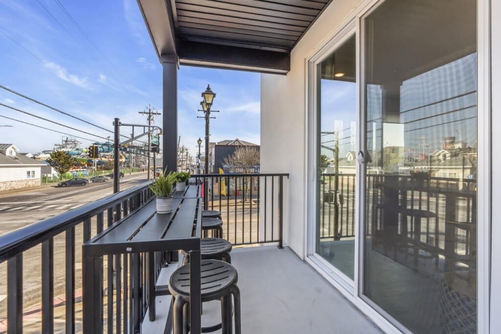 Balcony view of a shore property featuring a black table with stools, potted plant, and nearby street with traffic, reflecting the coastal environment of Jersey Shore real estate.