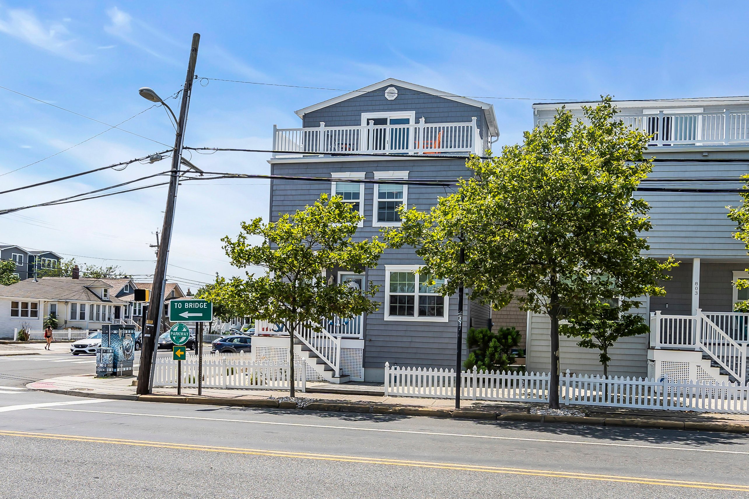 Gray beach house with balconies, surrounded by trees and a white picket fence, near street sign directing to the bridge.