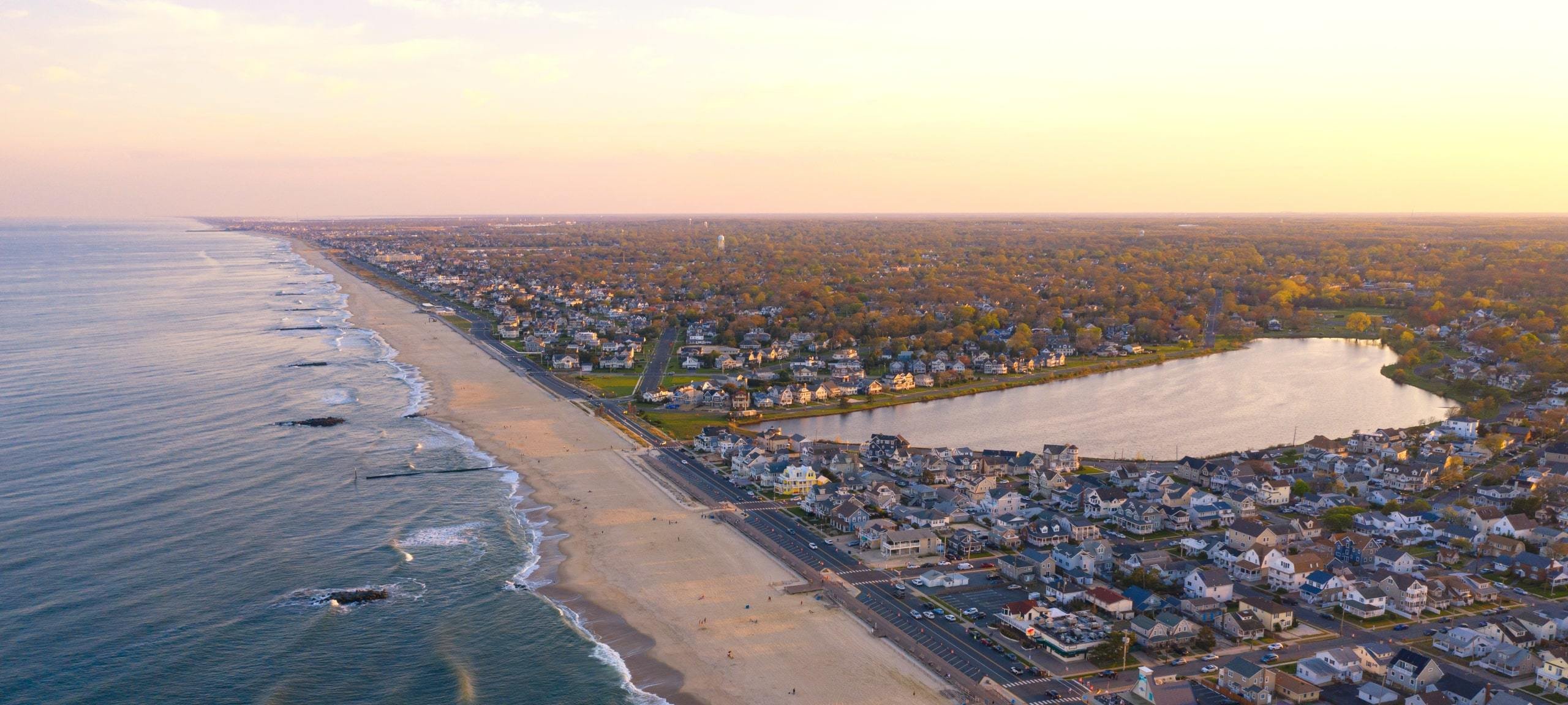 Aerial view of a sandy beach along the Jersey Shore, with minimal beachgoers, ocean waves, and a road lined with parked cars.