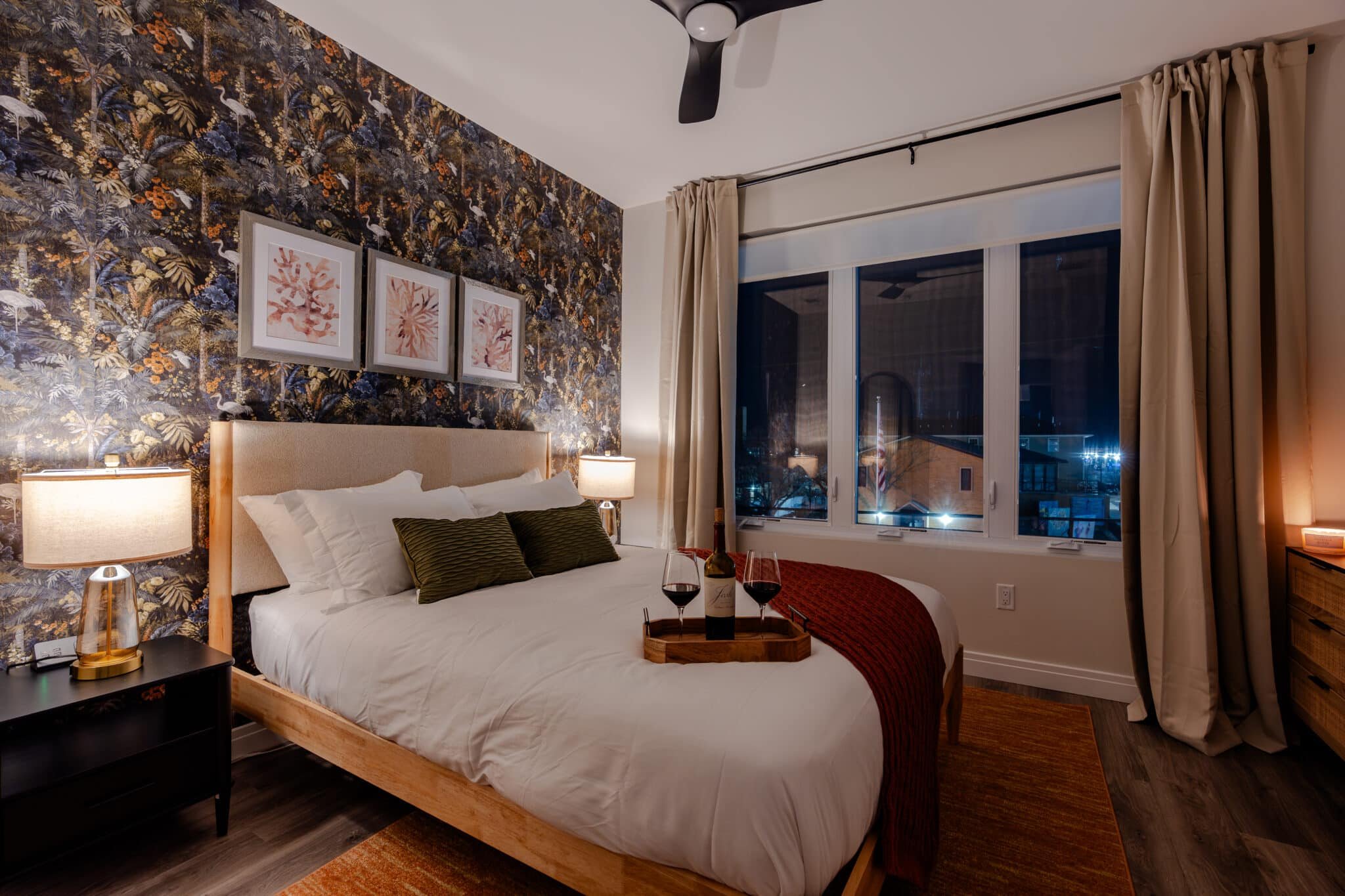 Cozy bedroom with modern decor, featuring a bed with decorative pillows, framed floral art on the wall, bedside lamps, and a wine tray.