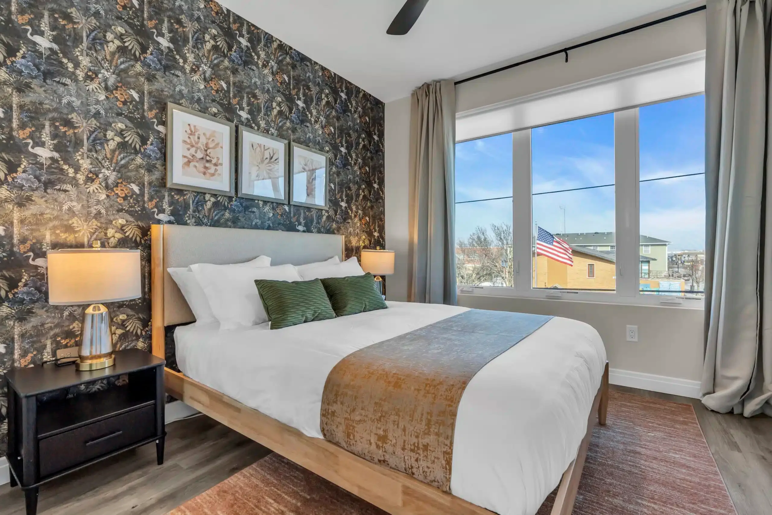 Cozy bedroom featuring a stylish bed with green pillows, decorative wall art, and large windows showcasing a view with an American flag, ideal for a Jersey Shore vacation rental.