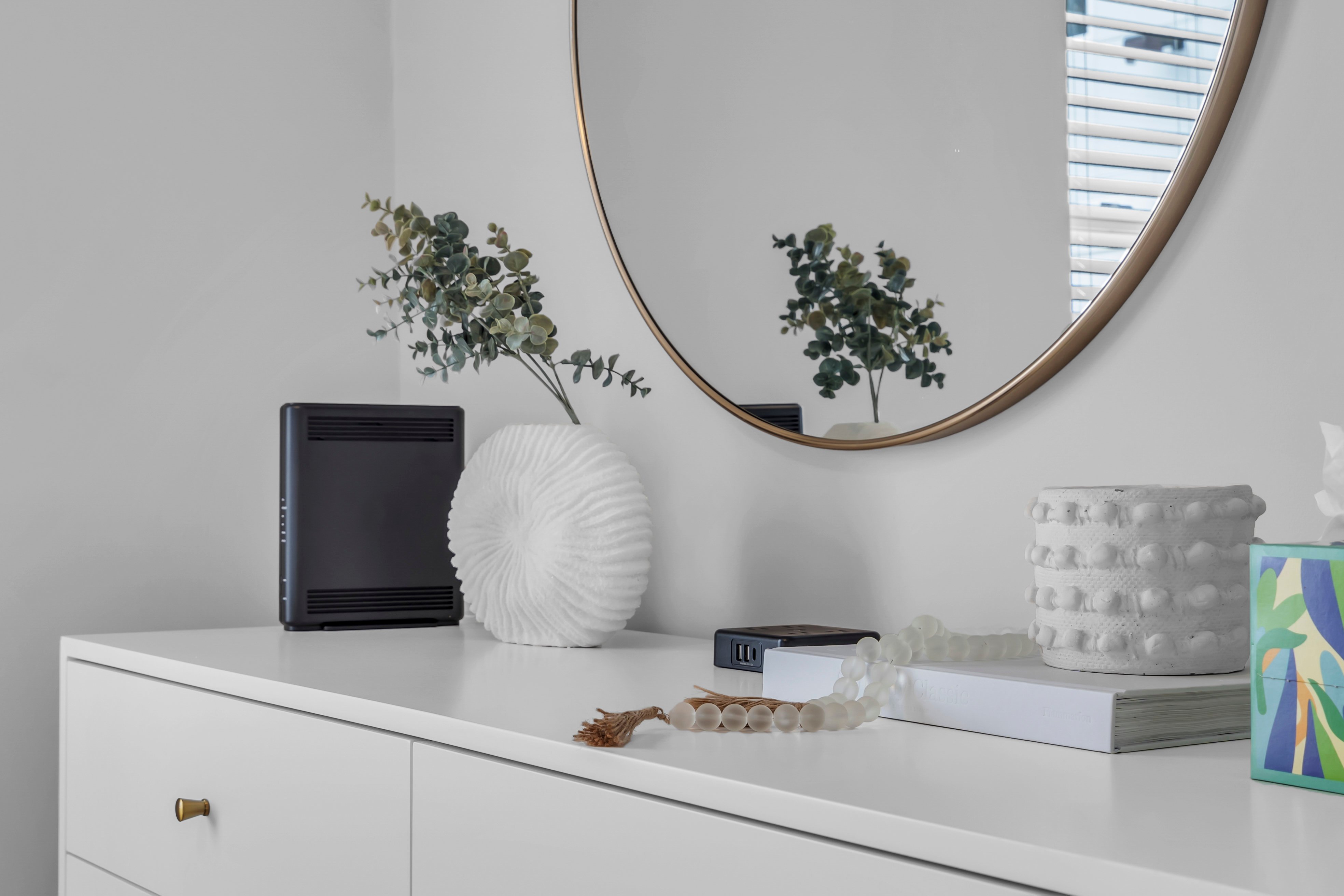 Modern coastal decor arrangement featuring a white dresser, decorative vase, greenery, and a round mirror, reflecting a serene beach house aesthetic.