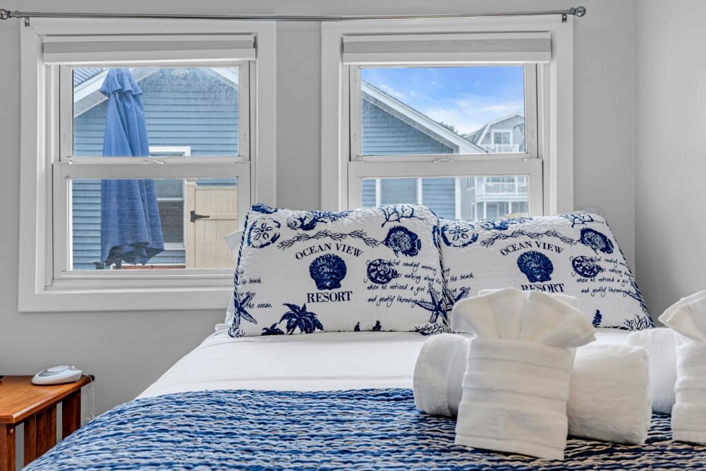 Cozy bedroom with ocean view pillows, white towels, and blue patterned bedding, showcasing a bright window view of the Jersey Shore vacation rental.