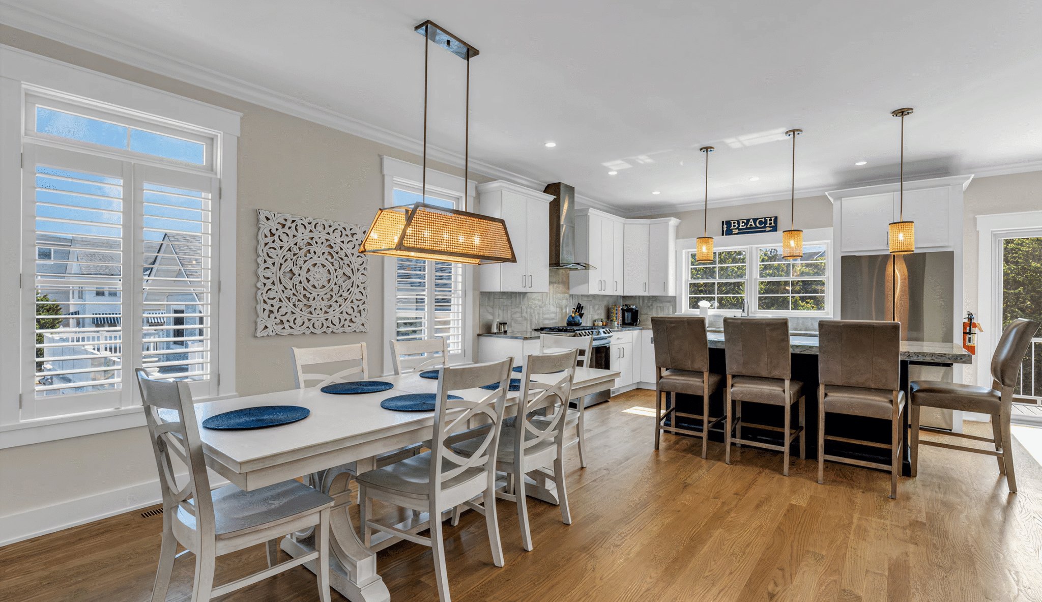 Modern beach house kitchen and dining area with white furniture, blue placemats, and large windows.
