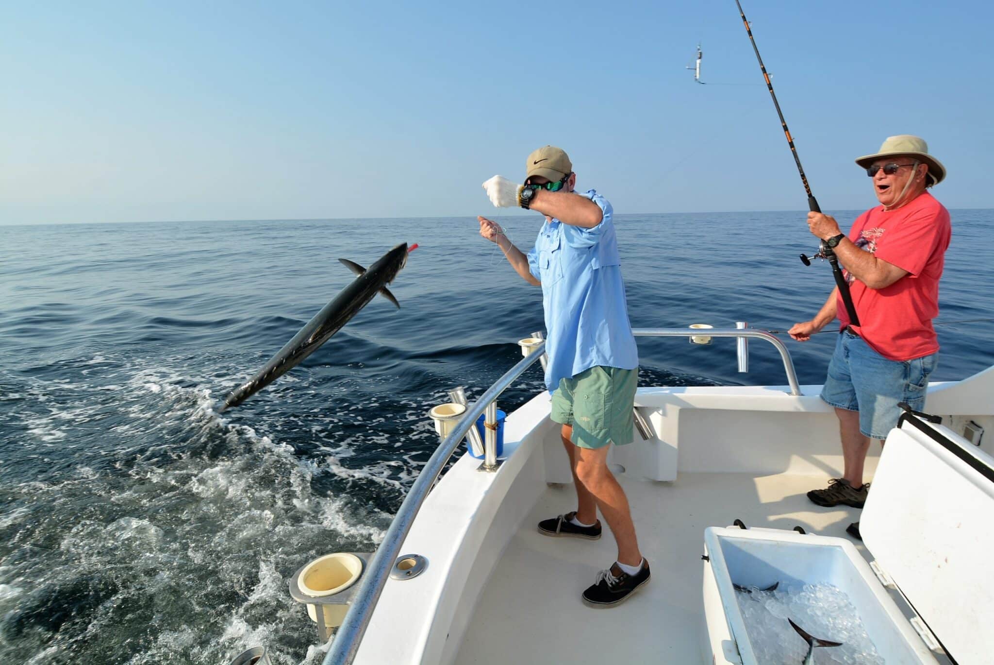Two fishermen on a boat at sea, one catching a kingfish leaping out of the water, highlighting the fishing experience off the Jersey Shore.