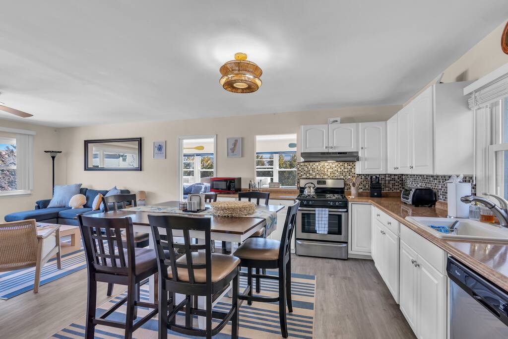 Modern beach rental kitchen and dining area featuring a spacious table, bar stools, stainless steel appliances, and a cozy living space with a blue sofa, ideal for maximizing guest experience and rental income.