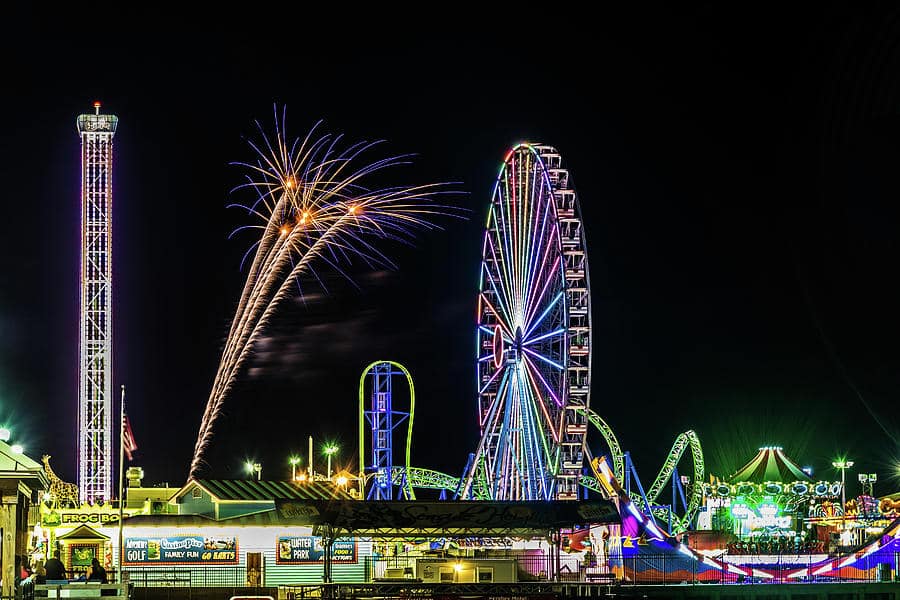 Fireworks illuminating the night sky above a vibrant Jersey Shore amusement park, featuring a colorful Ferris wheel and various rides, capturing the festive atmosphere of July 4th celebrations.