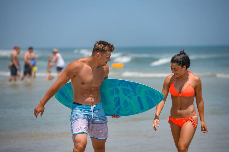 Young couple walking on the beach, smiling and enjoying the sun, with a surfboard, representing family fun and relaxation at Long Beach Island.