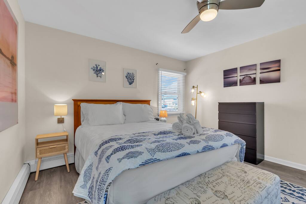 Cozy beach rental bedroom with ocean-themed decor, queen bed, bedside lamp, and dresser, emphasizing relaxing coastal atmosphere ideal for vacationers.
