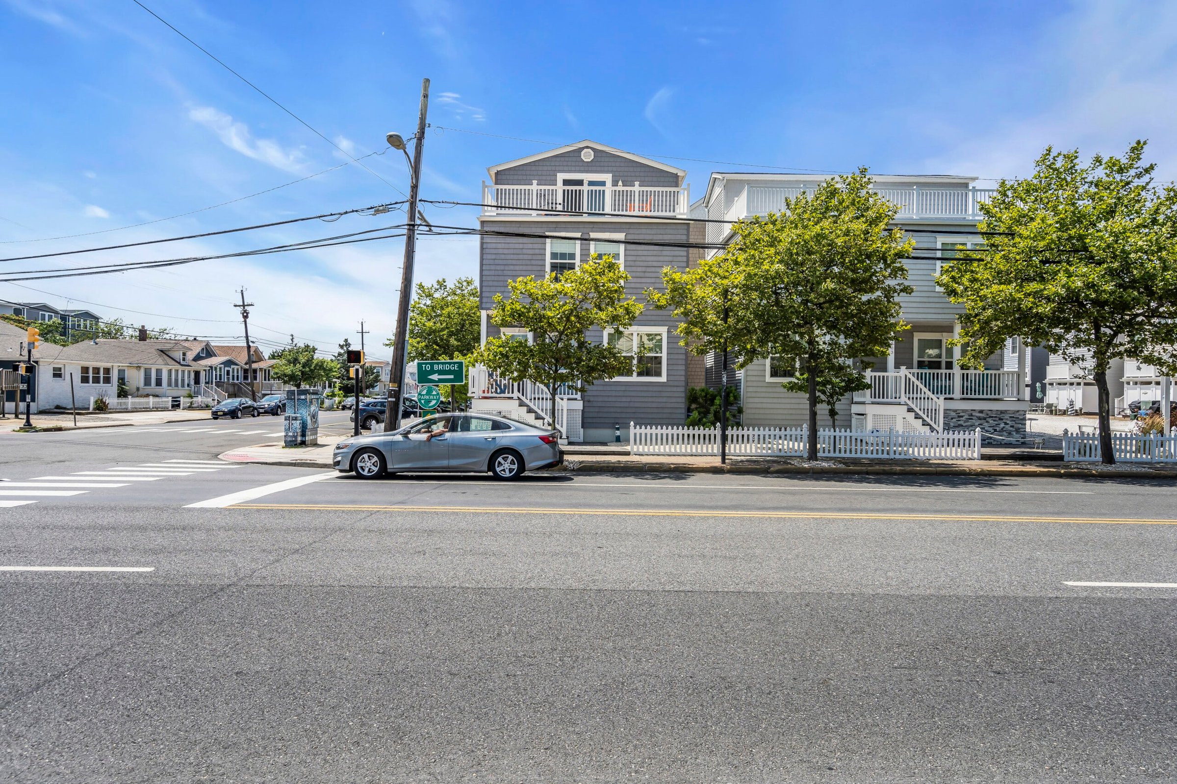 Coastal street view in Seaside Heights, NJ, featuring Breezy Beach Stays rental property, trees lining the sidewalk, and directional sign to the bridge.