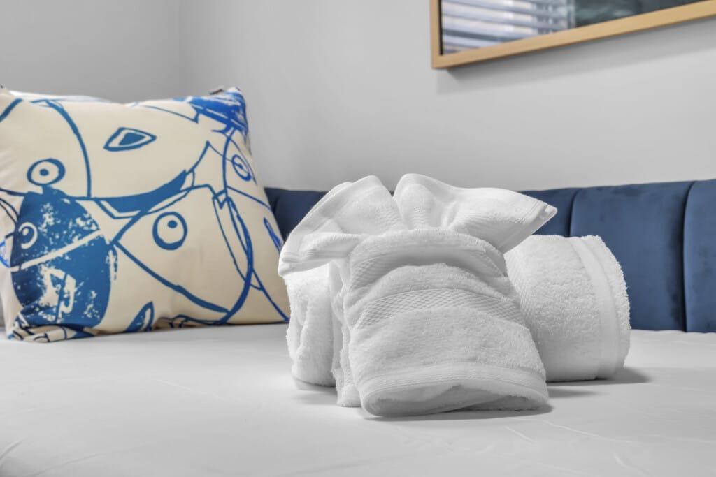 White towels neatly arranged on a bed with a decorative blue and white pillow, showcasing a beach-themed aesthetic suitable for shore home rentals.