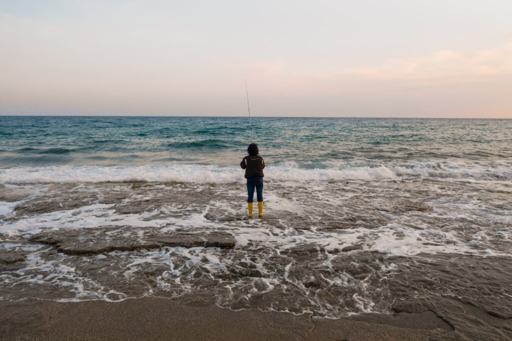 Person fishing at the shoreline in yellow waders, with ocean waves and a serene sky, representing fall fishing opportunities along the Jersey Shore.
