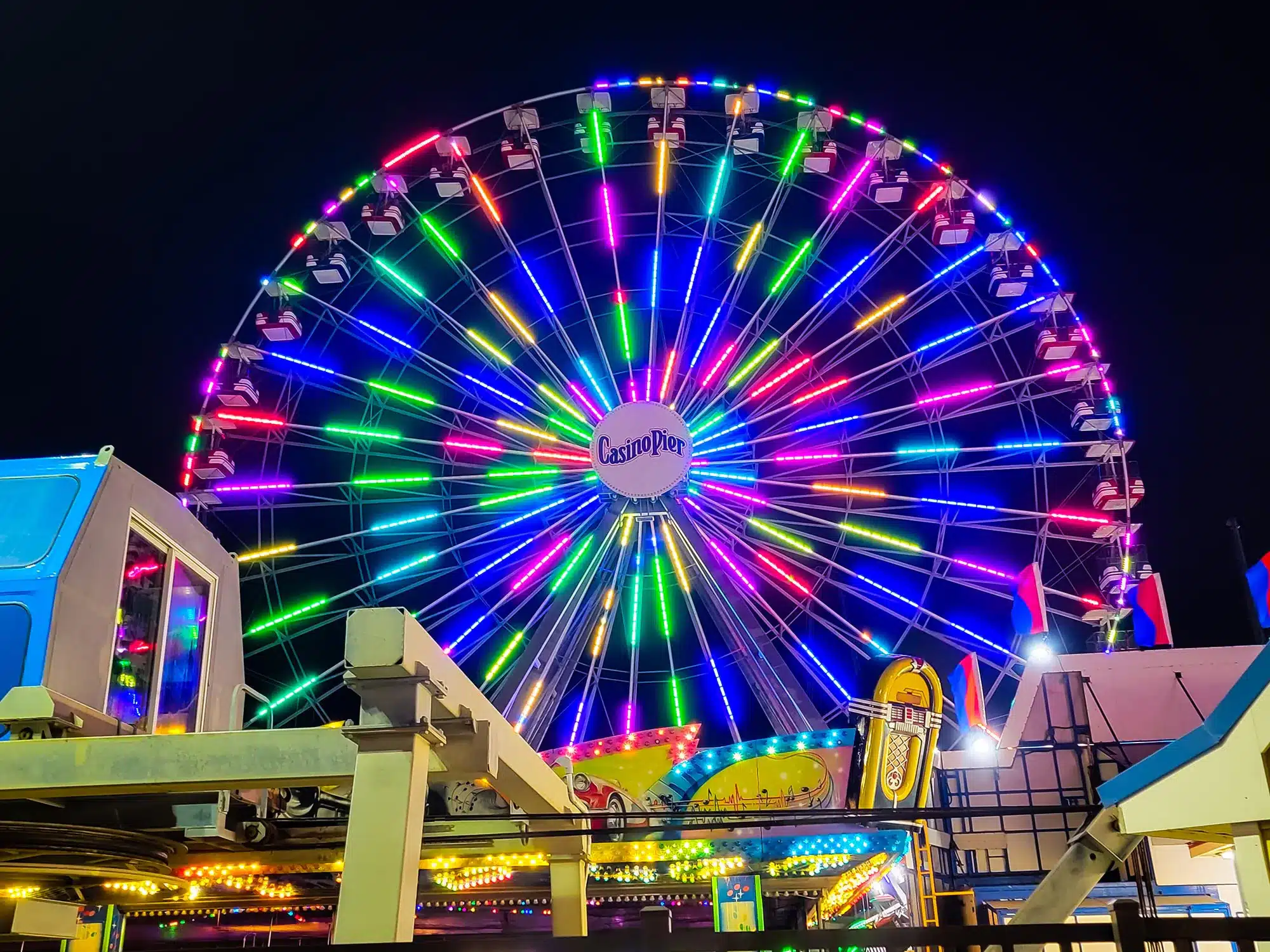 Colorful illuminated Ferris wheel at Casino Pier, surrounded by carnival attractions, showcasing vibrant lights against a night sky, highlighting Seaside Heights amusement park experience.