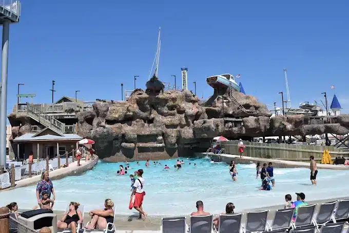 Breakwater Beach Waterpark featuring a large wave pool, families enjoying water attractions, and a rocky play structure with slides against a clear blue sky.