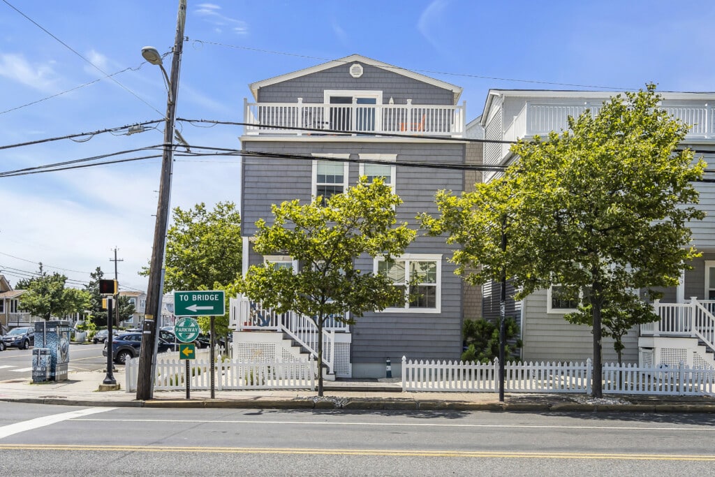 Coastal home exterior with gray siding, white trim, and balconies, surrounded by greenery and street signs indicating directions to the bridge and parkway, reflecting Jersey Shore property appeal.