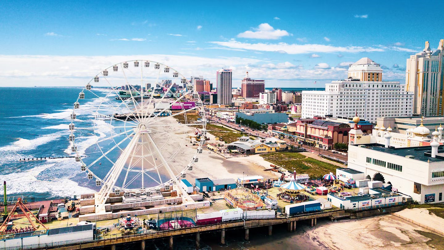 Aerial view of a vibrant boardwalk featuring a large Ferris wheel, beach attractions, and the Atlantic City skyline under a clear blue sky, highlighting popular summer activities at the Jersey Shore.