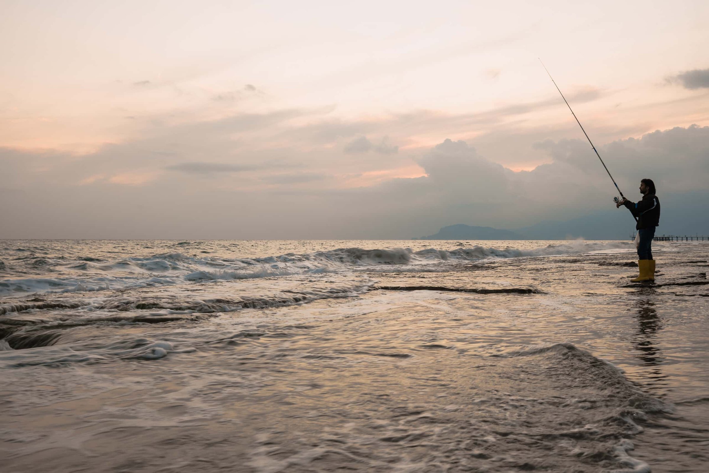 Man fishing on the Jersey Shore at sunset, wearing yellow waders, with ocean waves and a cloudy sky in the background, highlighting fall fishing opportunities.