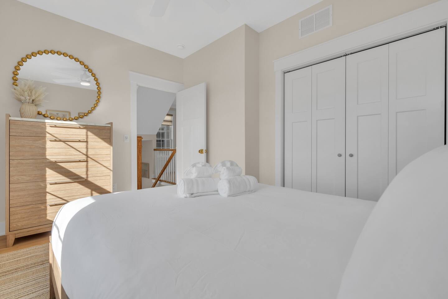 Cozy bedroom featuring a neatly made bed with rolled towels, wooden dresser, round mirror, and light-colored walls, illustrating inviting vacation rental accommodations at the Jersey Shore.