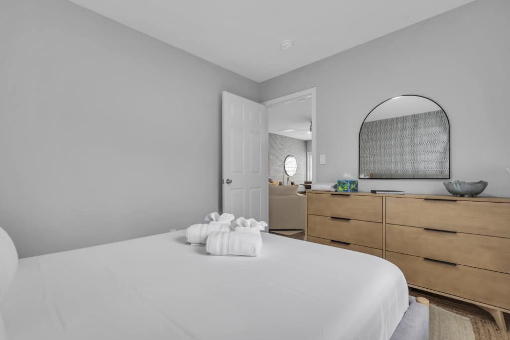 Modern bedroom with a neatly made bed featuring rolled towels, light gray walls, a wooden dresser, and a round mirror reflecting a cozy living area, highlighting the inviting atmosphere of Jersey Shore properties.