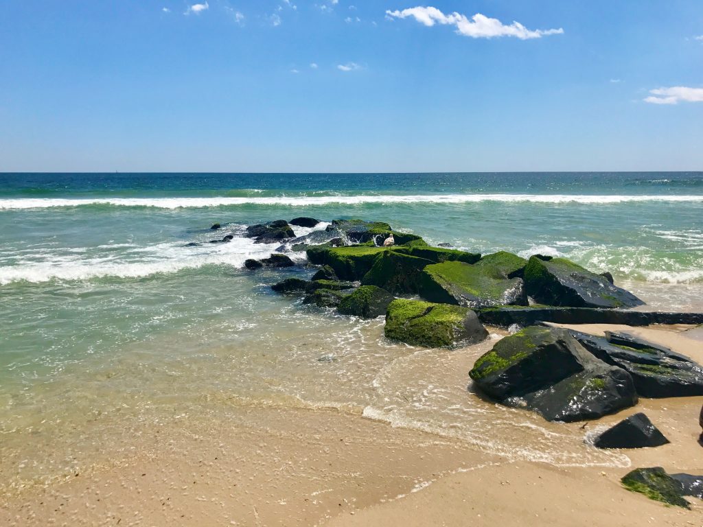 Rocky jetty covered in green moss, with gentle ocean waves and clear blue skies, highlighting the tranquil beach atmosphere of Lavallette, Jersey Shore.