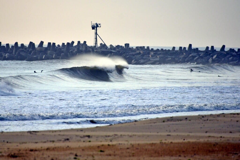 Surfing waves at Manasquan Inlet Beach with jetty in the background, showcasing ideal surf conditions and experienced surfers.