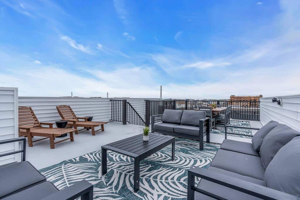 Rooftop deck featuring modern outdoor furniture, including gray sofas and wooden lounge chairs, with a view of the Jersey Shore skyline under a bright blue sky, ideal for vacation relaxation and gatherings.