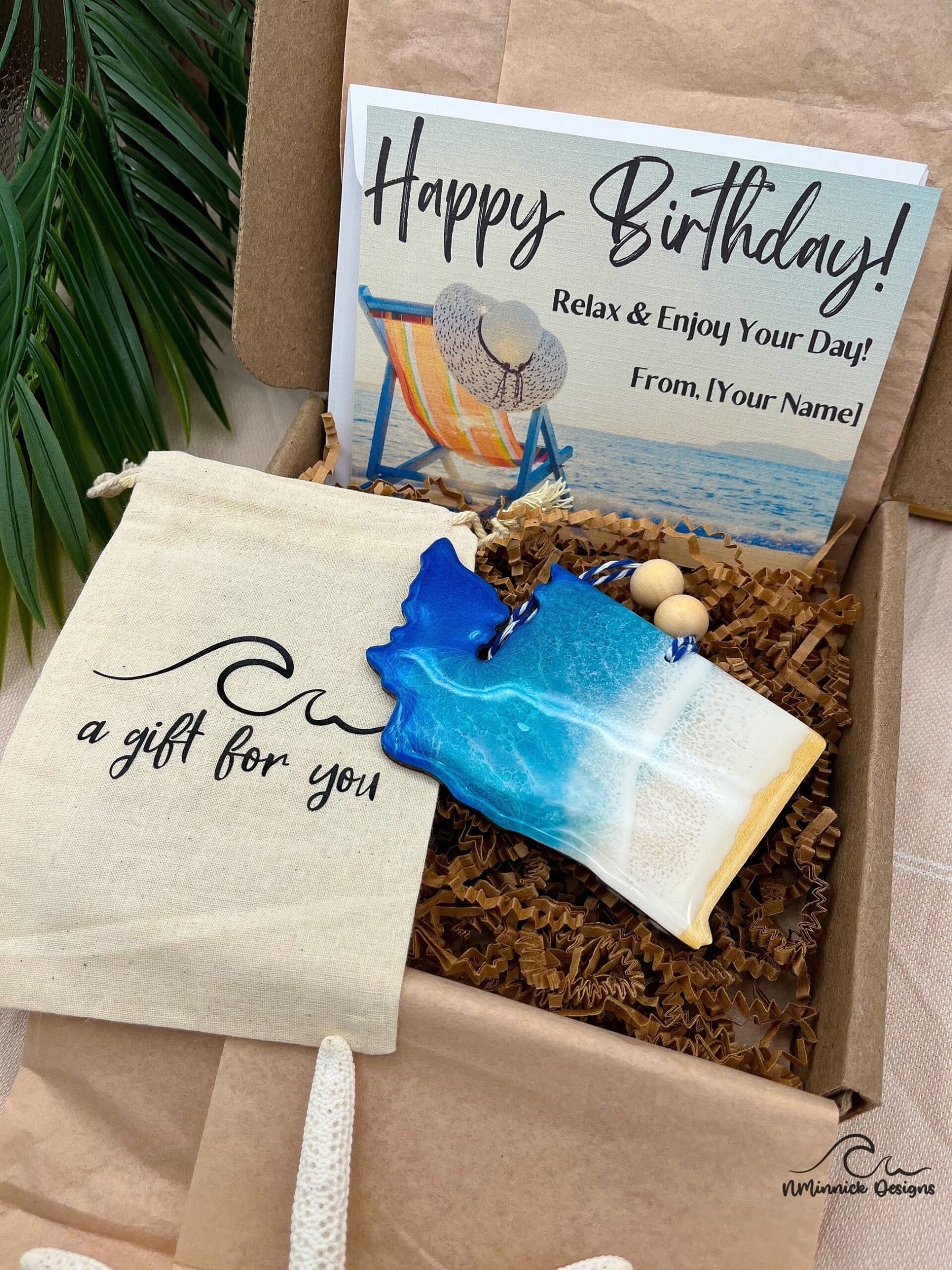 Gift box featuring a beach-themed birthday card, a decorative blue and white ocean-inspired ornament, and a canvas pouch labeled "a gift for you," surrounded by crinkle paper and tropical foliage, emphasizing unique souvenirs at the Jersey Shore.