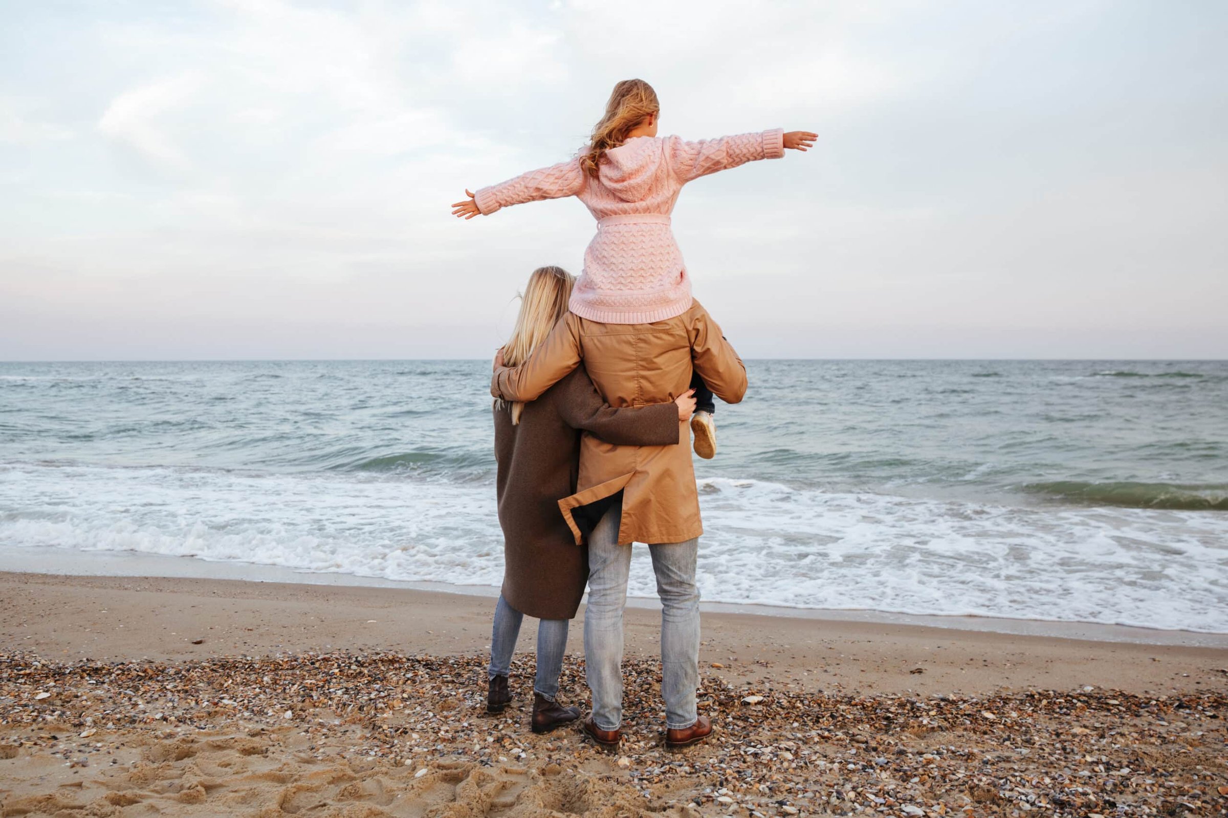 Back view of a smiling family at the beach, with a little girl in a pink sweater playfully perched on her father's shoulders, enjoying a family-friendly