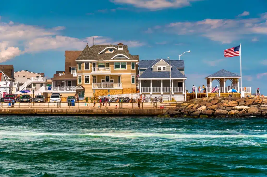 Colorful beach houses along the Lavallette waterfront with American flag, people enjoying the beach, and scenic ocean waves, highlighting family-friendly vacation rentals at Breezy Beach Stays.
