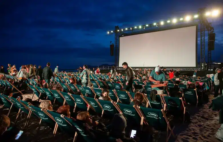 Beach movie screening setup with audience gathering, sandy setting, large screen and chairs under evening sky.