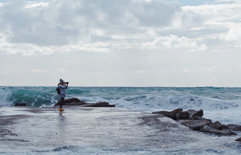 Woman photographing ocean waves from a rocky jetty, capturing the dynamic coastal scenery ideal for fall fishing along the Jersey Shore.