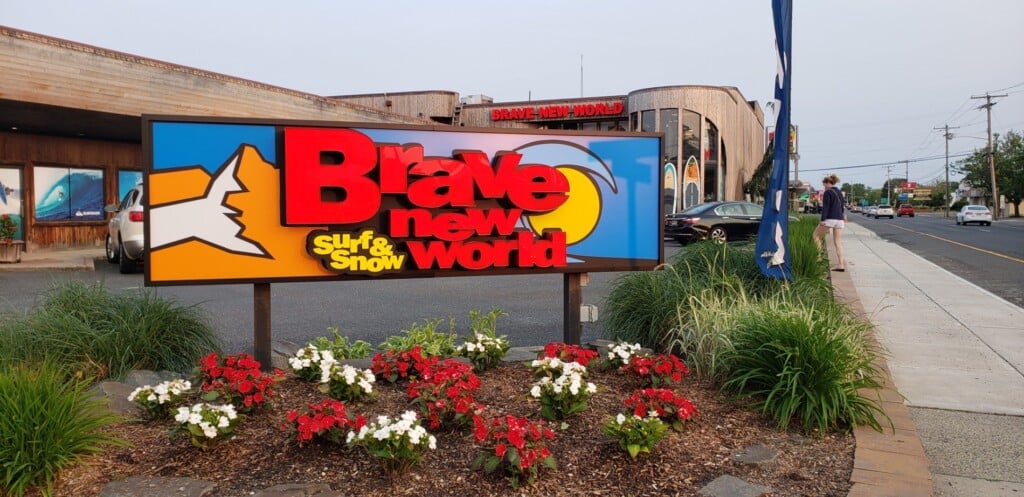 Brave New World Surf Shop sign with vibrant colors, surf and snow theme, surrounded by flowers, located at the Jersey Shore, reflecting the local surf culture and community.