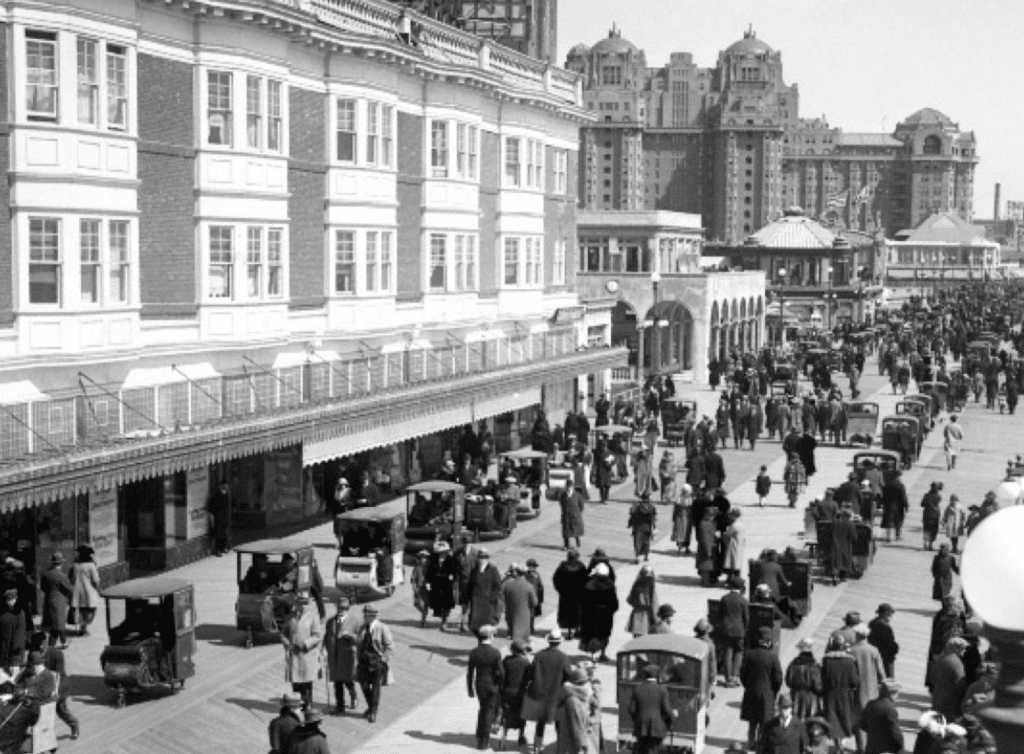 Crowd bustling on Atlantic City Boardwalk in 1923, featuring vintage architecture, shops, and early automobiles, reflecting the rise of beach culture and tourism.