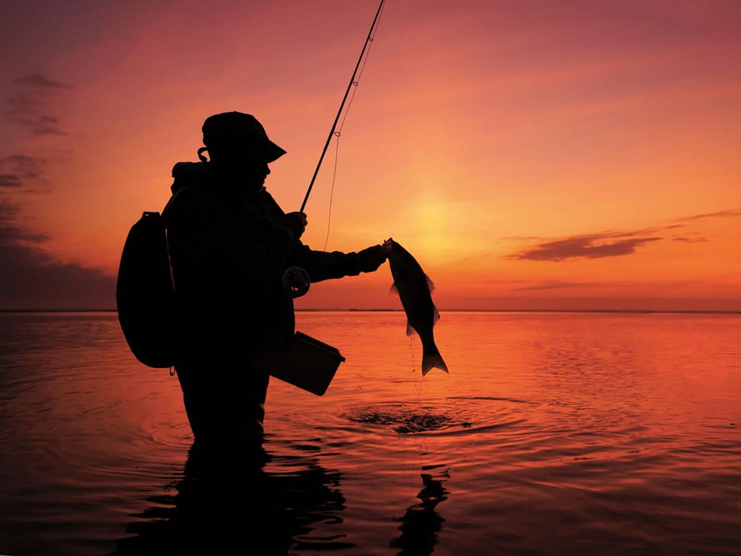 Silhouette of a fisherman holding a caught striped bass against a vibrant sunset over Barnegat Bay, illustrating fishing activities near Seaside Heights.